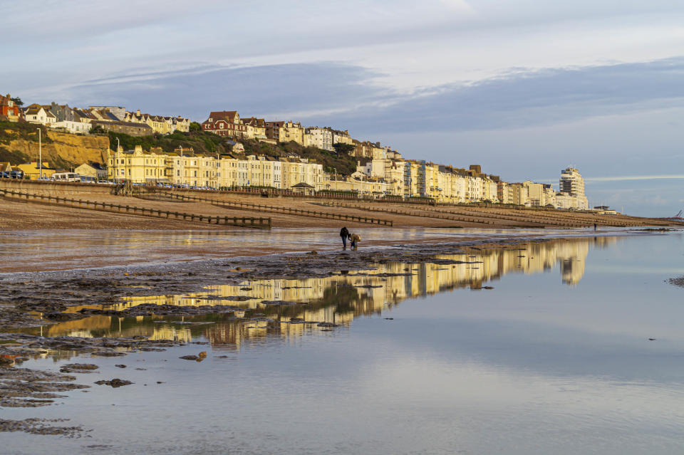 A view of the seafront of West Marina in St Leonards-on-Sea, East Sussex, taken from the beach at low tide. The buildings are reflected in the calm sea and sand and the setting sun has made the building glow.