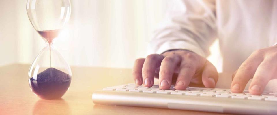 Man's hands typing on computer keyboard next to a hourglass. Concept of time management, business schedule and deadline, for background, website banner, promotional materials, advertising.