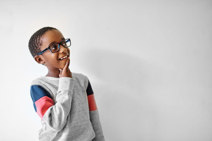 A young child wearing glasses and a sweater with patterned sleeves stands against a plain background, smiling thoughtfully with a finger on their chin