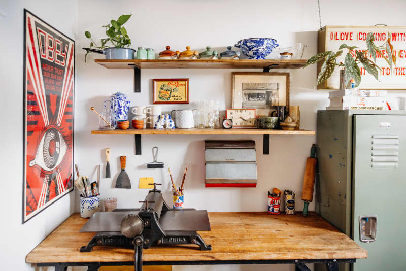 Wooden work table and wall shelves decorated with vintage items are seen in white room.