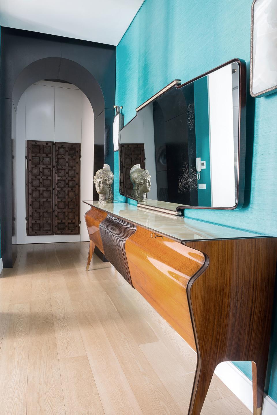 In this hallway leading to the kitchen, a midcentury rosewood sideboard by Osvaldo Borsani stands against a wall covered in turquoise silk wallpaper, which is hung with a set of backlit onyx and cast-bronze sconces designed by Salvagni. Toward the back, behind a black-stained oak arch, is a wardrobe with antique Tibetan doors.