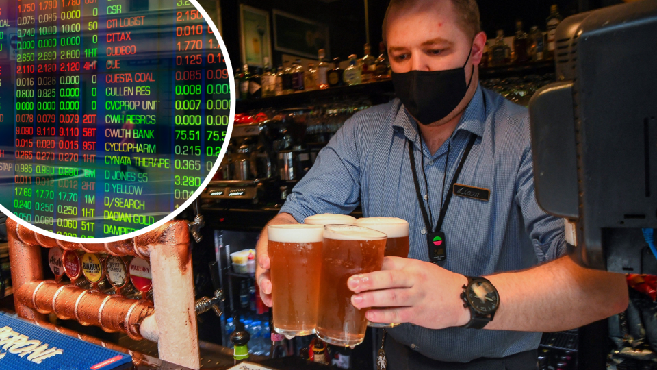 A person serves four freshly poured beers from behind the bar of a pub while wearing a face mask and the ASX board showing company prices changes in the corner.