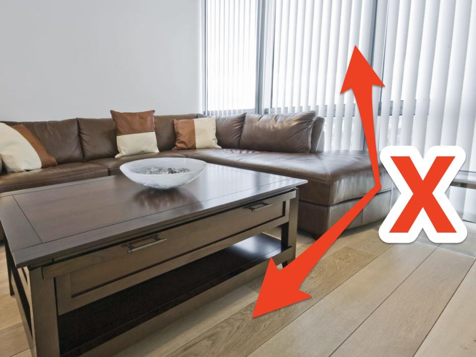 A living room with a brown couch and coffee table with bare floors and vertical blinds with red arrows and X pointing to blinds and floor