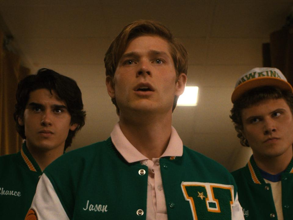 jason, played by mason dye, standing looking surprised. there are other members of the hawkins basketball team, wearing lettermen jackets, standing behind him