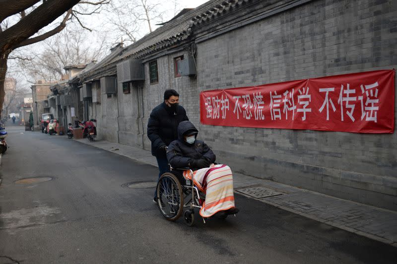 Man pushes a man on wheelchair past a propaganda banner on a wall in a hutong in Beijing