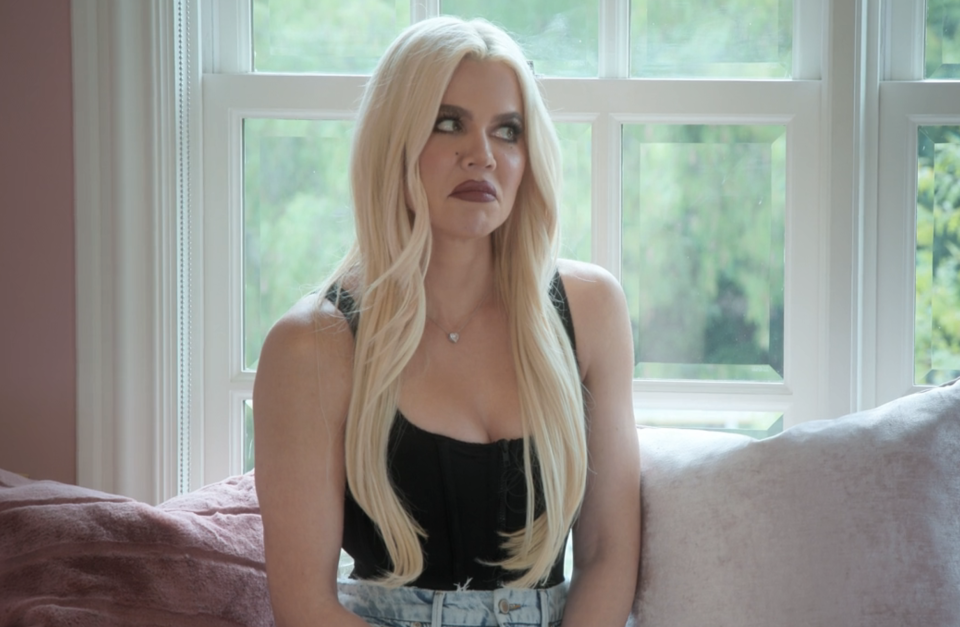 Khloé Kardashian sits on a couch wearing a black sleeveless top and light blue jeans. The image is from a TV show episode