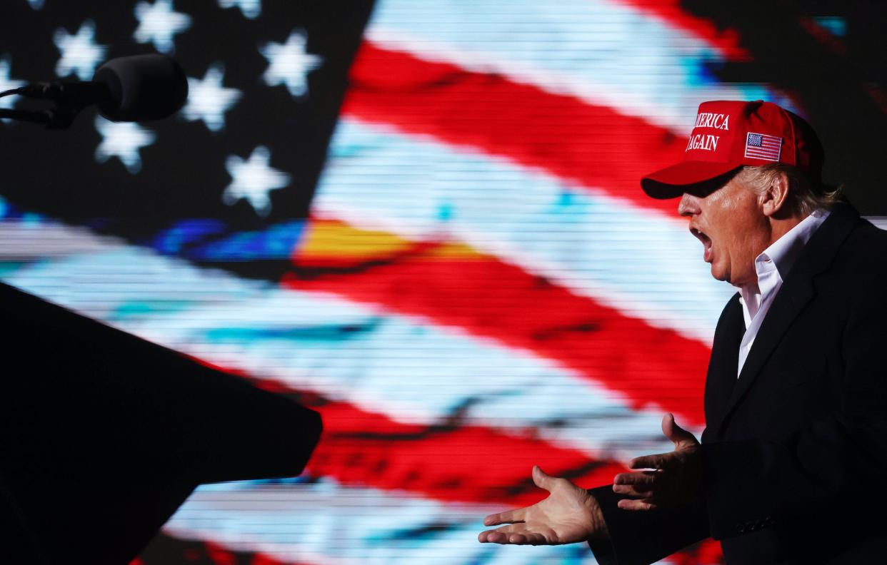 Donald Trump yells in front of an American flag while wearing a MAGA hat.