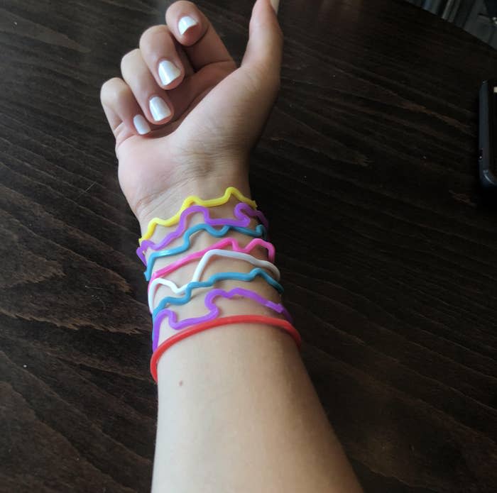 A person's arm is covered with several colorful, wavy rubber bands. The person has white nail polish and is resting their arm on a dark wooden surface