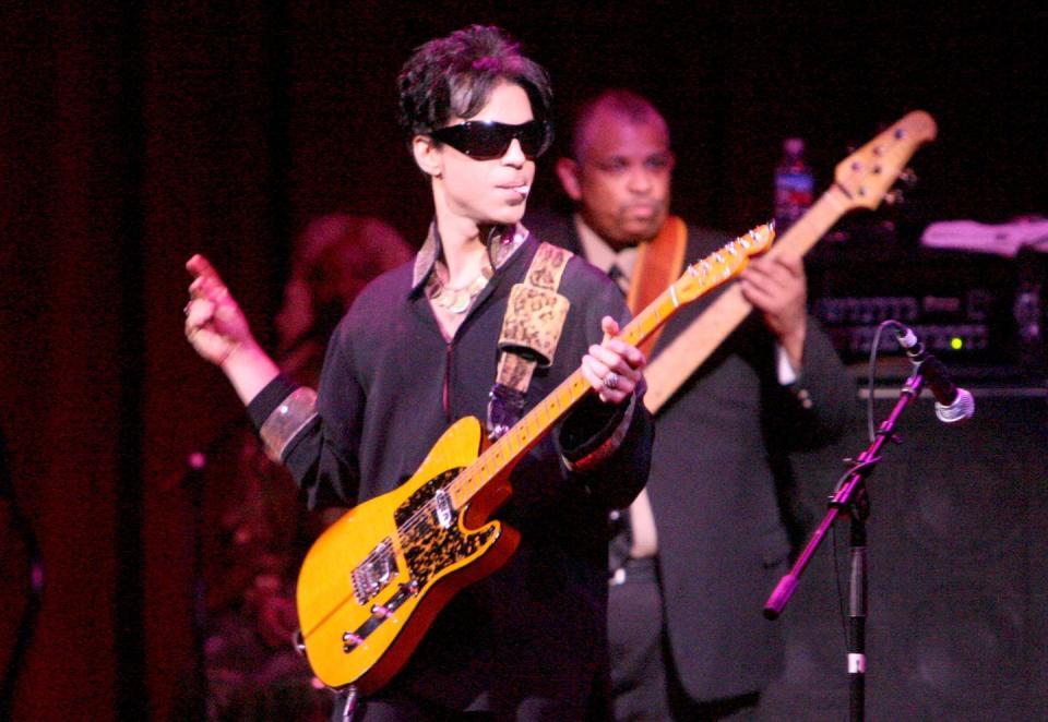 prince plays a guitar and looks to the right, he wears sunglasses and a dark shirt