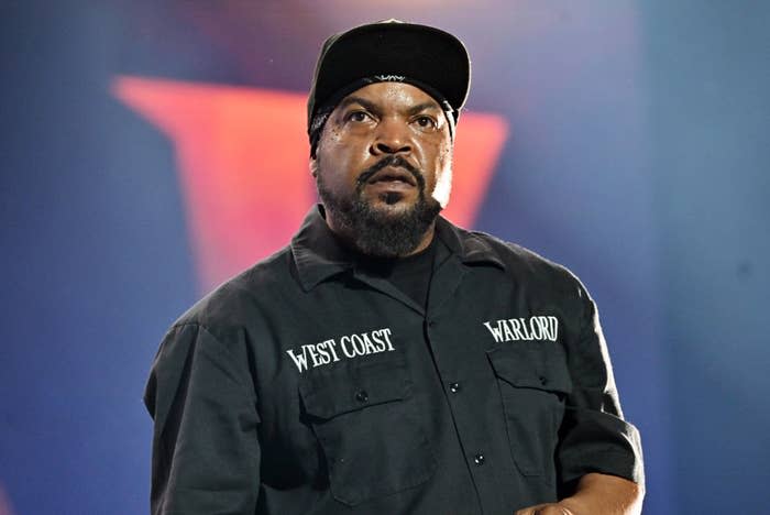 Ice Cube wearing a black shirt with "West Coast" and "Warlord" text, and a cap, standing on stage