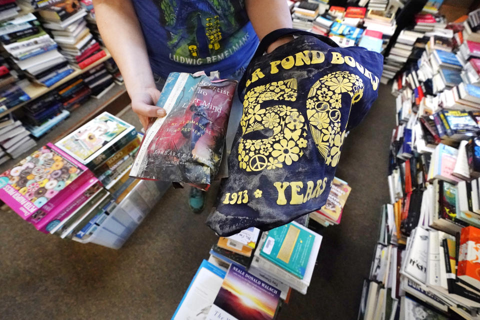 Bookstore manager Cora Kelly holds up a flood damaged book and shirt while inventory of undamaged books are stacked at Bear Pond Books, Tuesday, Aug. 1, 2023, in Montpelier, Vt. The mostly gutted shops, restaurants and businesses that lend downtown Montpelier its charm are considering where and how to rebuild in an era when extreme weather is occurring more often. Vermont's flooding was just one of several major flood events around the globe this summer that scientists have said are becoming more likely due to climate change. (AP Photo/Charles Krupa)