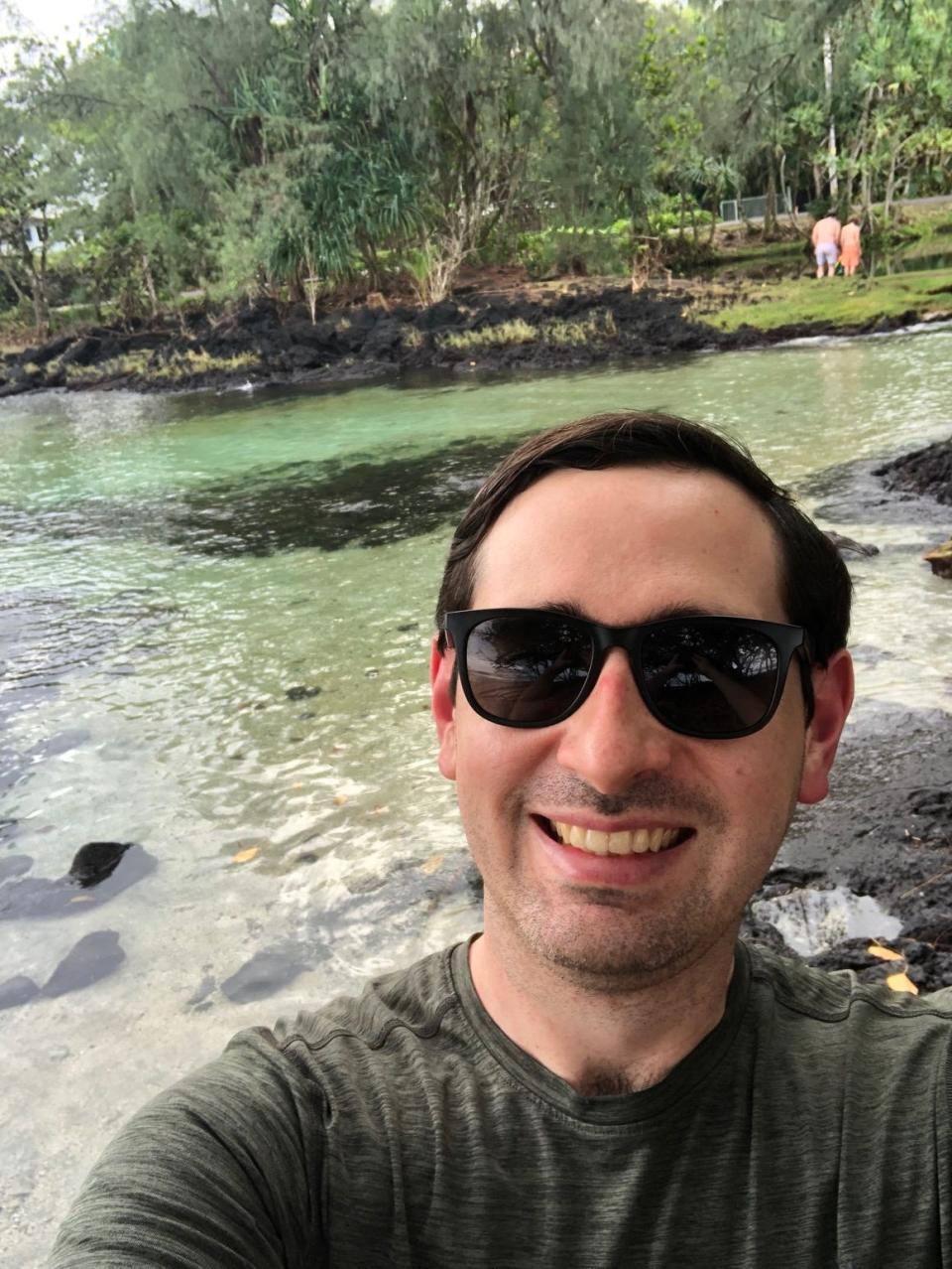 Reporter Alex Bitter wearing sunglasses and a dark green shirt standing near the water at a beach outside Hilo, Hawaii
