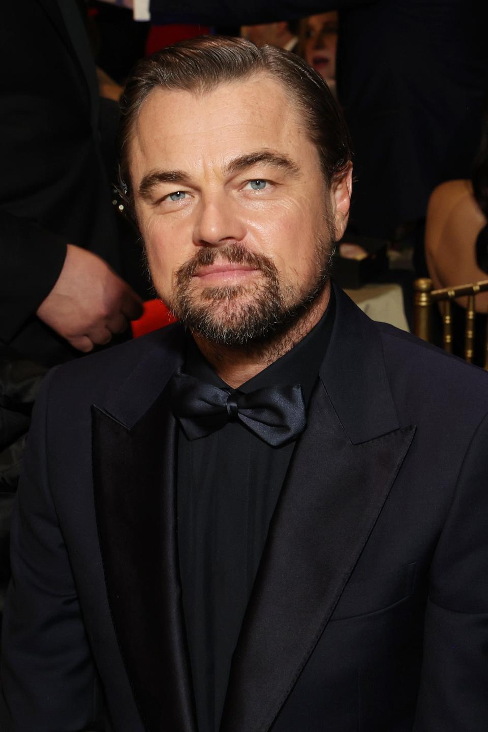 Leonardo DiCaprio wearing a black tuxedo and bow tie at an event. He has a beard and styled hair