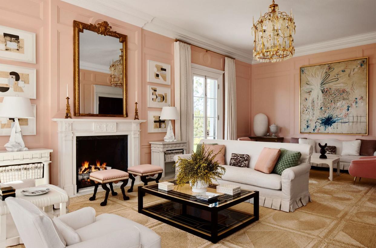 a seashell pink living room has two slipcovered sofas, upholstered chairs, cocktail table, fireplace with a large gilt antique mirror above it and flanked by tables with lamps, artworks, a patterned sisal rug