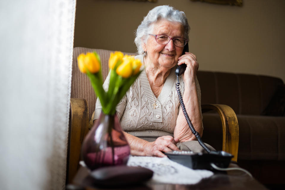 Old woman with gray white hair and glasses sitting in her armchair in her home and talking on wired phone as Australian government introduces smart phone coronavirus applications