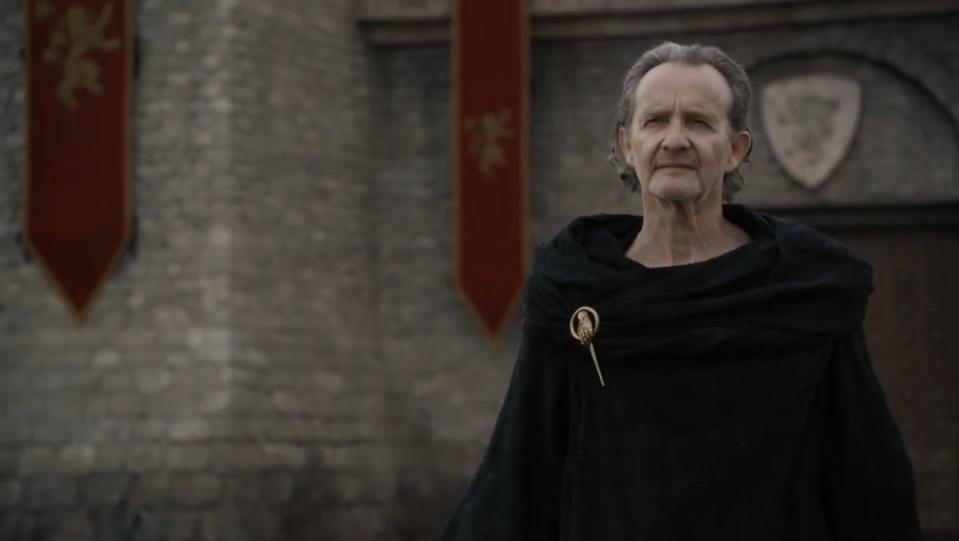 A skinny older man in black robes walks outside the gates of a castle.