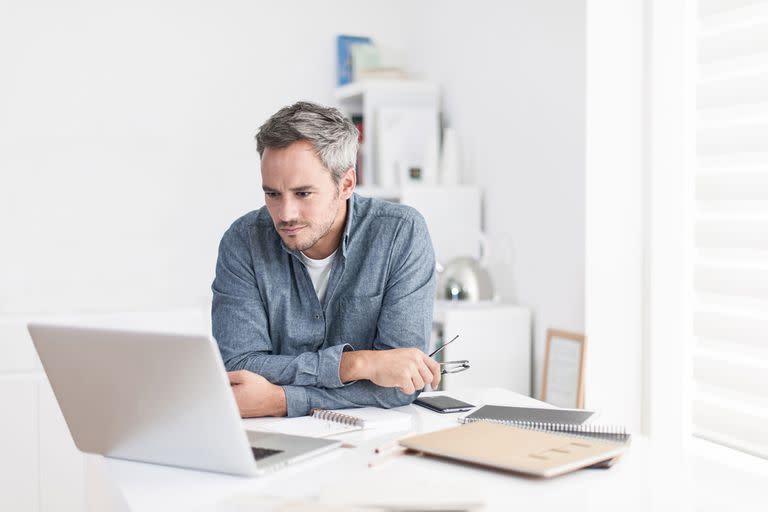 Portrait of a nice smiling grey hair man with beard holding his glasses, working at home on some project, he is sitting at a white table looking at his laptop in front of him. Focus on the man