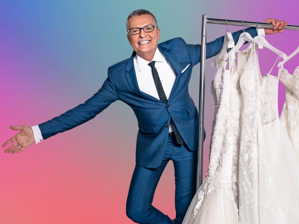 A photo of Randy Fenoli hanging off of a rack of wedding dresses in front of a rainbow backdrop.