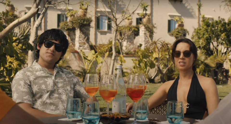 A man and a woman sit at an outdoor table with drinks and glassware in front of them. The man wears a floral shirt and sunglasses; the woman wears a black halter top and sunglasses