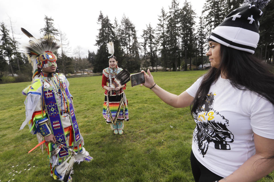 In this photo taken Saturday, April 4, 2020, Wakiyan Cuny, 16, left, and his sister Wicahpi Cuny, 14, Dakota and Lakota tribal members, wear ceremonial clothing while they are filmed by their mother Tera Baker during a live streamed powwow from a park near their home in Puyallup, Wash. The largest powwows in the country have been canceled or postponed amid the spread of the coronavirus. Tribal members have found a new outlet online with the Social Distance Powwow. They're sharing videos of colorful displays of culture and tradition that are at their essence meant to uplift people during difficult times. (AP Photo/Elaine Thompson)