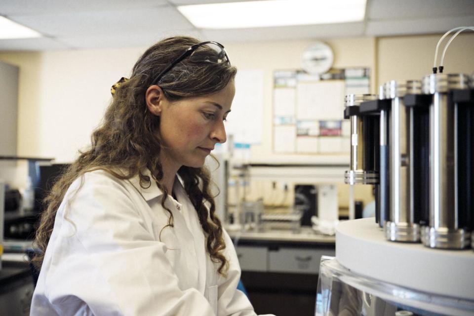 Μια γυναίκα με λευκό εργαστηριακό παλτό στέκεται δίπλα σε επιστημονικό εξοπλισμό.