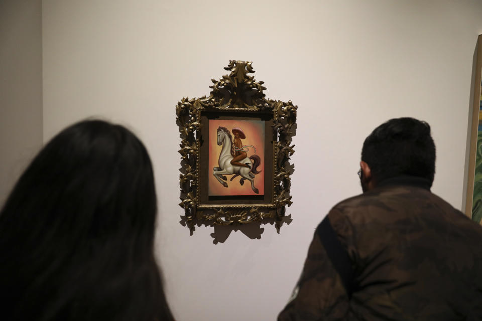 People watch a painting showing 1910-17 Mexican revolutionary hero Emiliano Zapata nude, wearing high heels and a pink, broad-brimmed hat, straddling a horse, in Mexico City, Tuesday, Dec. 10, 2019. The work by Fabian Chairez is part of an exhibit about Zapata in one of Mexico City's premiere arts venues, the Fine Arts Palace. (AP Photo/Eduardo Verdugo)