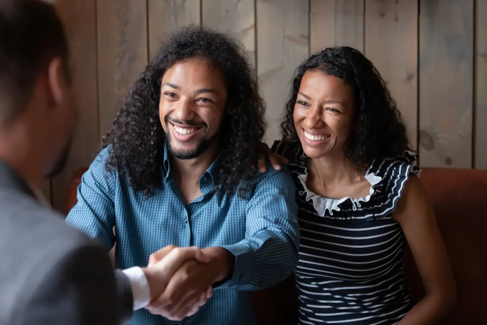 Happy emotional african american family couple shaking hands with professional broker or Real Estate Agent, starting meeting in modern cafe or office. Smiling biracial clients making agreement with agent.