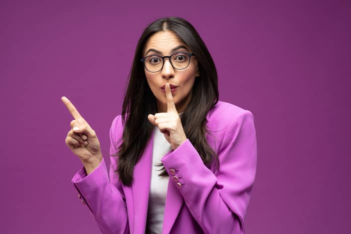 Woman in a blazer gestures to be quiet, looking surprised, with both index fingers raised