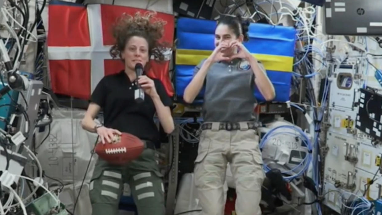  Two astronauts floating in a module. one holds a football. flags are behind them. 