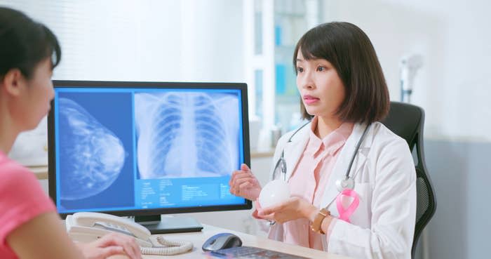 A doctor with a pink ribbon on her coat shows a breast model to a patient. X-rays are visible on a computer screen in the background