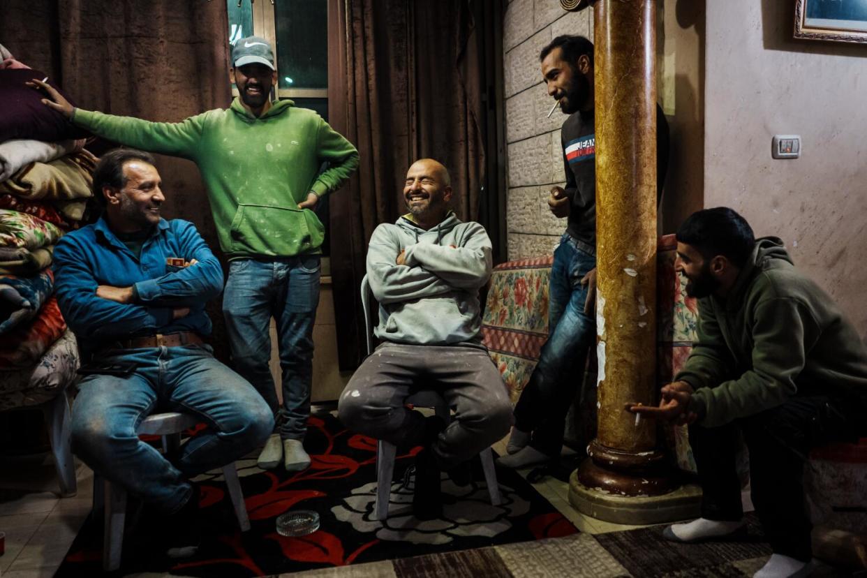 Five men laughing while gathered in a room, three sitting and two standing