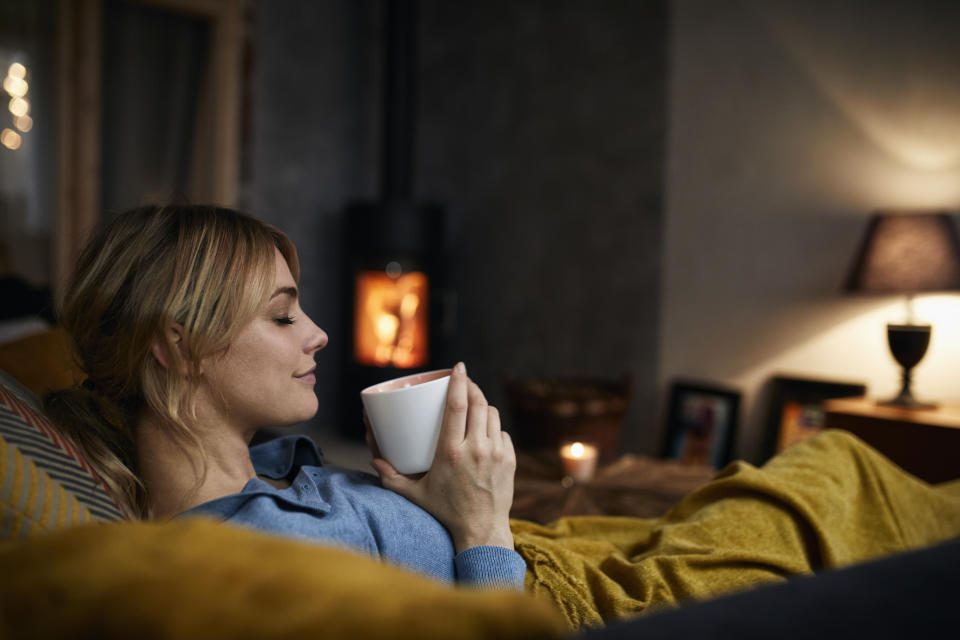 Frau sitzt mit Decke und Tasse im Wohnzimmer vor Kamin und Kerze