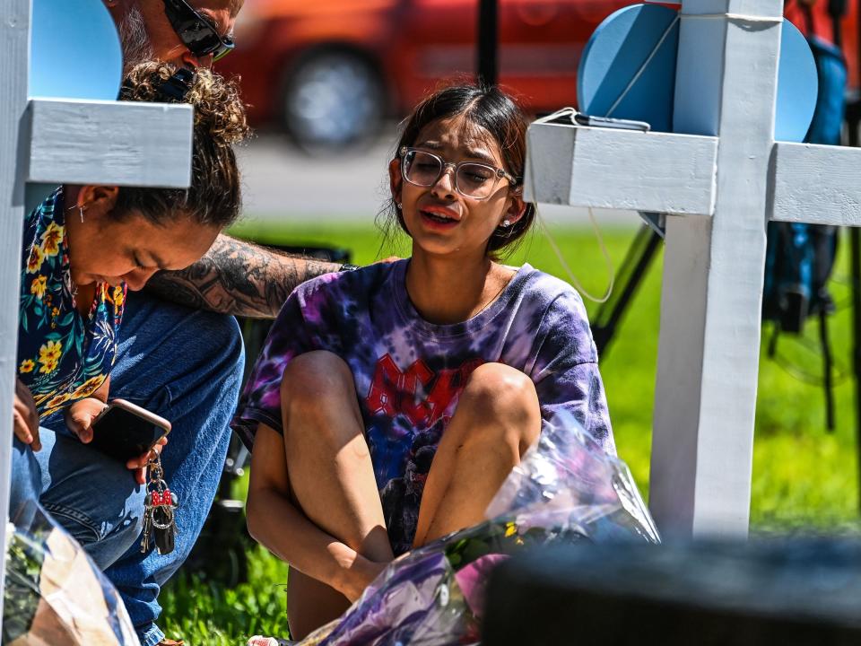 A girl sits in front of a cross holding her knees and crying.