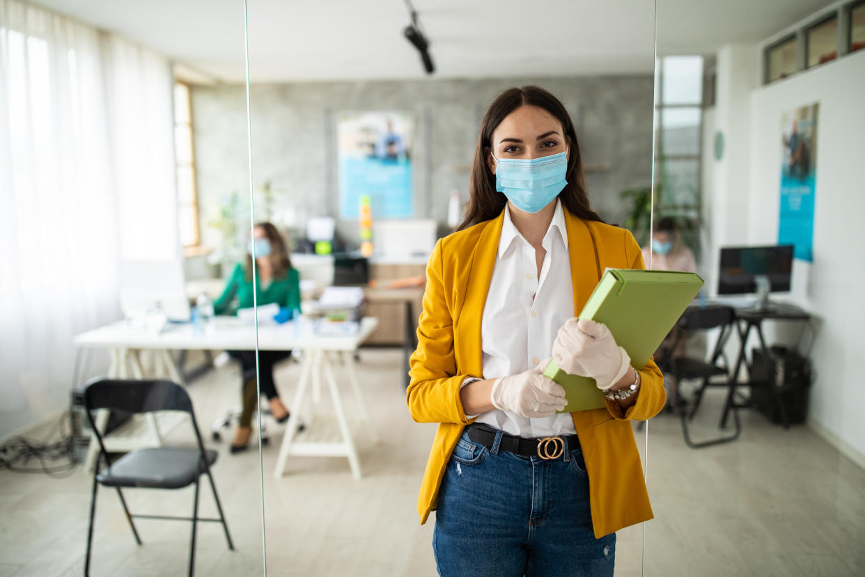 Portrait of female bank manager with protective face mask and surgical gloves in office, standing, looking at camera and smiling during covid-19 pandemic