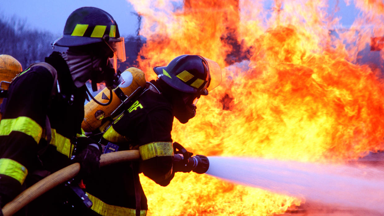  We see two firefighters in full gear holding a hose and blasting close by flames. 