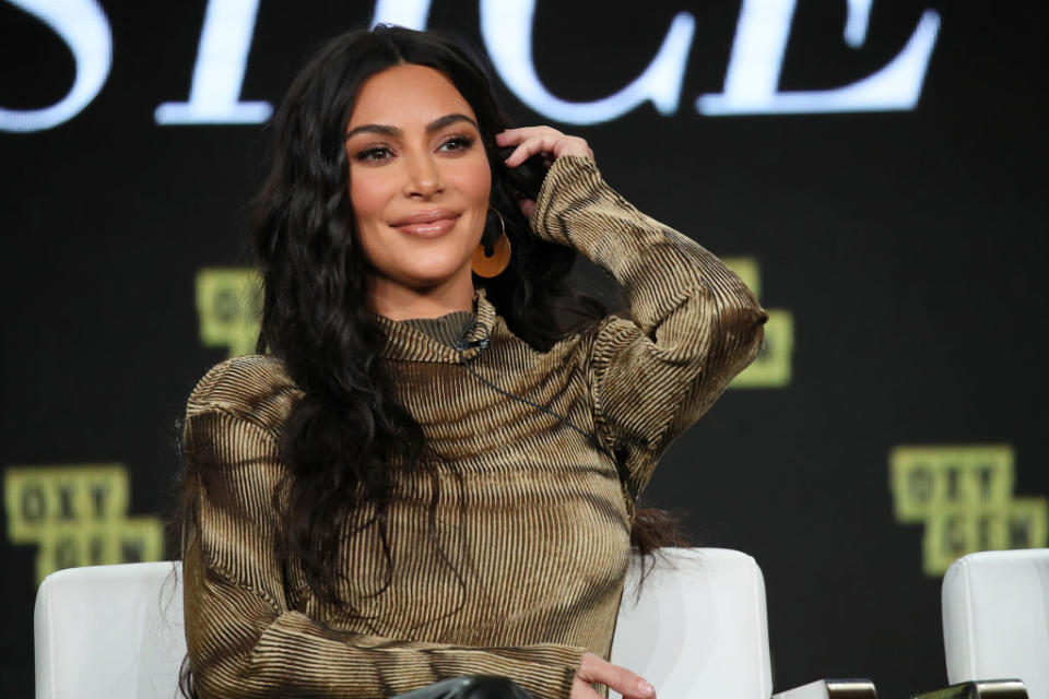 Kim Kardashian is seated, wearing a long-sleeve ribbed top, smiling, and touching her hair. A backdrop with indistinct text is behind her