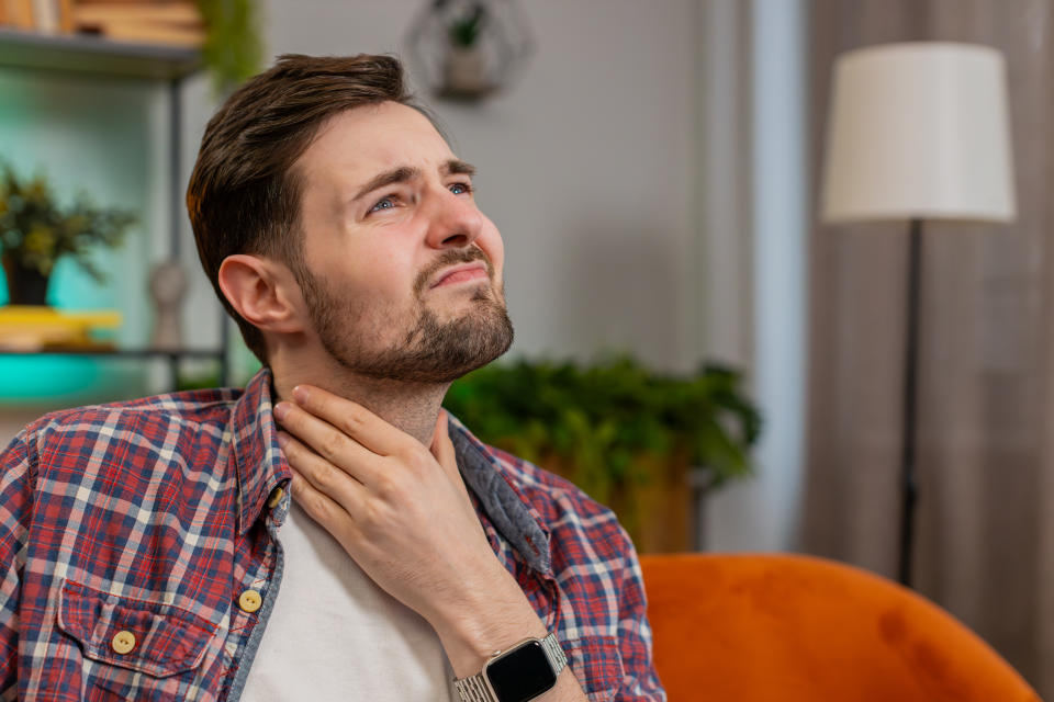 Upset Caucasian man in plaid shirt holds hands over sore throat. Painful neck and frowning, thyroid disorders, suffering sore throat, tonsils inflammation. Young guy sitting on sofa at home room