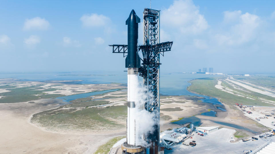 a black and white rocket stands next to a huge metallic launch tower, with the ocean in the background