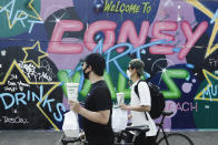 Cyclists wearing protective masks walk with food from Nathan's Famous past a graffiti art mural by Tats Cru toward the boardwalk of Coney Island Beach Friday, May 15, 2020, in the Brooklyn borough of New York. (AP Photo/Frank Franklin II)