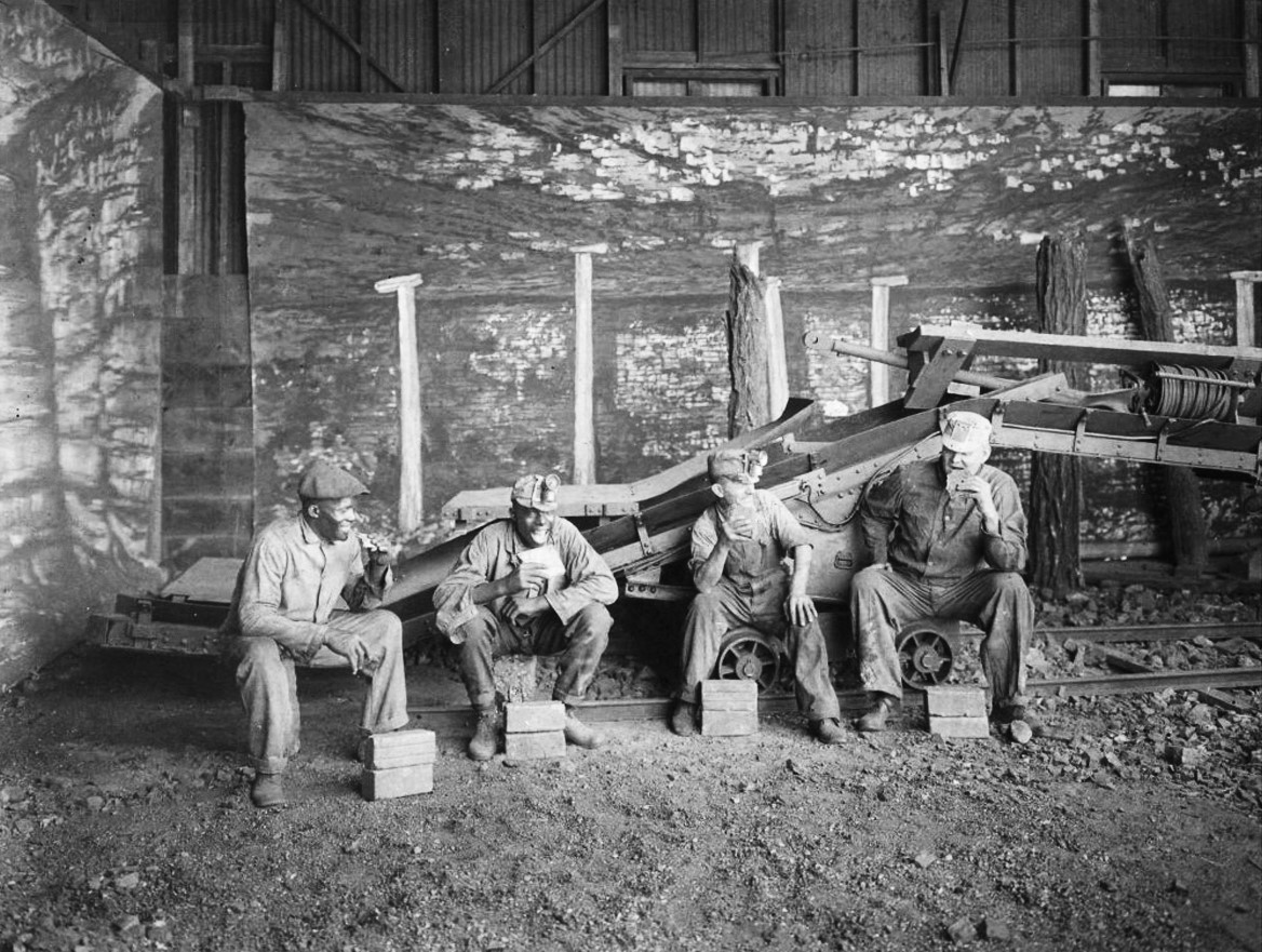 These "miners" are sitting in front of a coal loader made by the Jeffrey Manufacturing Company of Columbus. Corporate photographer and Grandview resident Fred Behmer often staged environments that would be similar to where Jeffrey Manufacturing equipment would be used for purposes of documentation and advertising. This 1918 photograph of a mine scene used a background painted to look like the interior of a coal mine. It was created inside the Jeffrey plant, with Jeffrey employees dressed like miners having a lunch break.