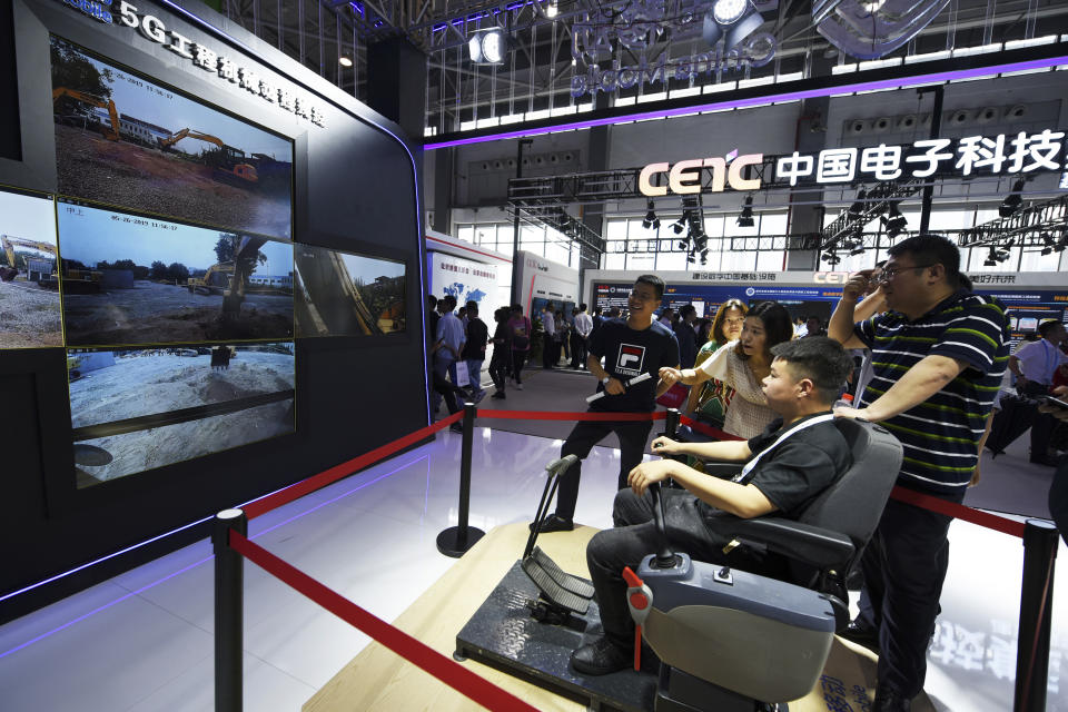 In this Sunday, May 26, 2019, photo released by Xinhua News Agency, a visitor operates an excavator which is shown on the TV screens by a remote control system via 5G technology at the China International Big Data Industry Expo 2019 in Guiyang, southwest China's Guizhou Province. China's latest media broadside in its trade war with the U.S. features an unlikely star: Trevor Noah of the Daily Show. The state broadcaster CCTV ran part of a segment from the show on Chinese equipment maker Huawei's dominance in 5G mobile technology in its lunchtime news show Monday, May 27. (Yang Wenbin/Xinhua via AP)