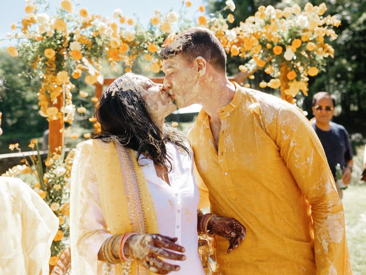 A bride and groom kiss in front of floral archway during their haldi ceremony.