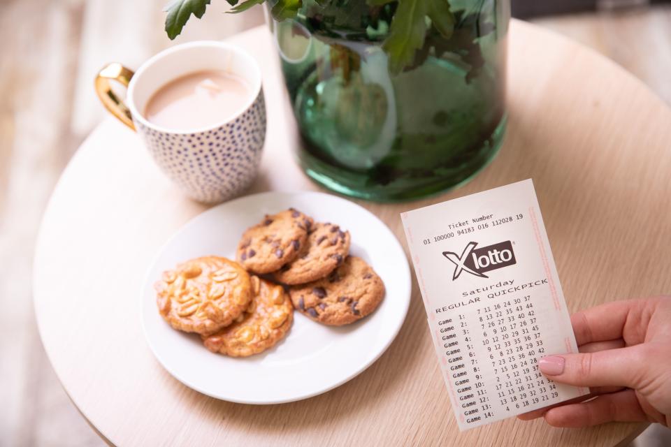 A woman holds a lottery ticket next to a cup of tea and biscuits. Source: The Lott