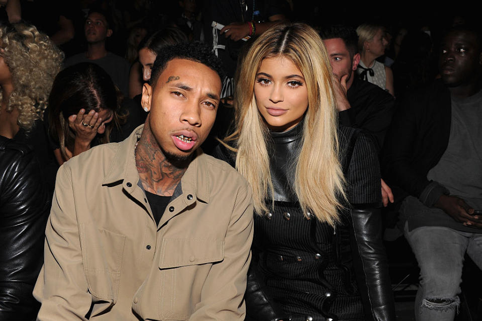 Tyga and Kylie Jenner at a public event, sitting close together. Tyga is in casual attire, while Kylie is wearing a stylish black outfit