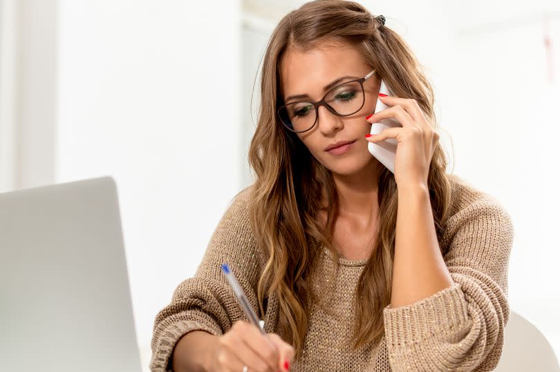 Getty generic. Young woman talking on mobile phone and writing notes while sitting at her desk with open laptop. Pretty caucasian female working in home office.