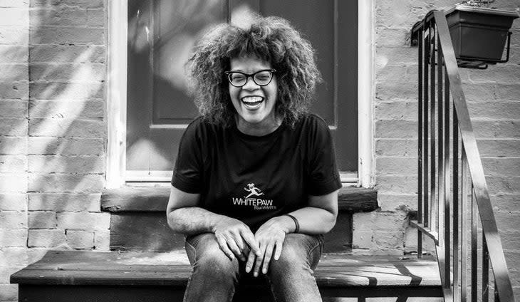 smiling woman wearing glasses and black t-shirt and jeans sitting on steps in front of a door