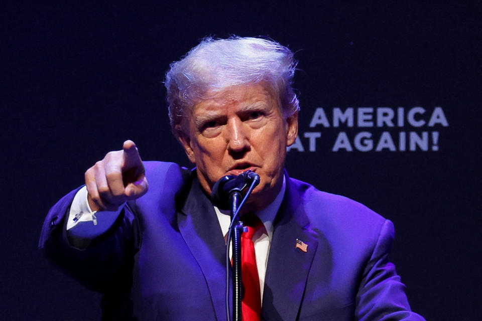 Donald Trump stands at a microphone and points toward the camera.