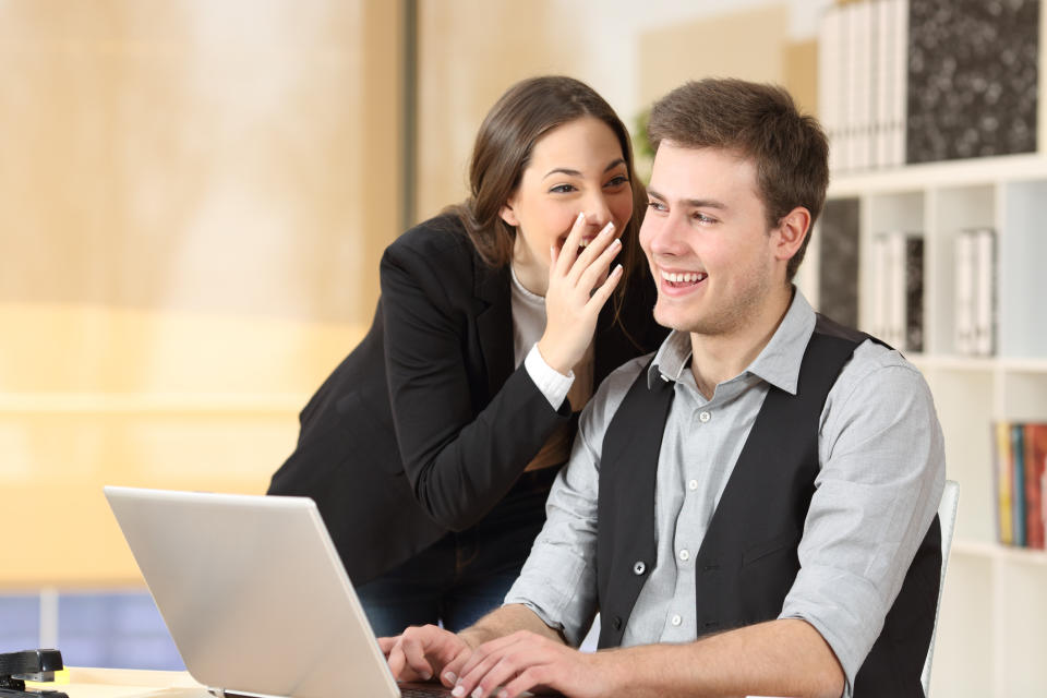 Young woman whispering into the ear of a male colleague. Both of them are smiling from ear to ear.