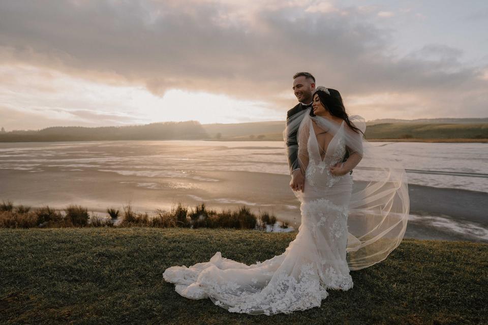 A bride and groom embrace on a grassy hill in front of the ocean.