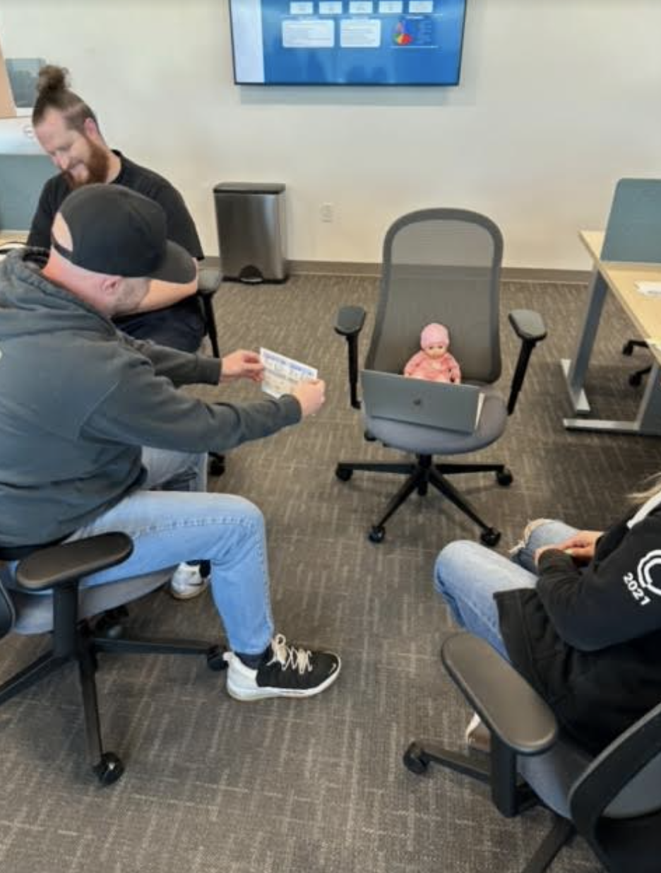 Four people are seated in an office. Three adults surround an infant in a baby seat on a swivel chair. One adult is pointing to a piece of paper